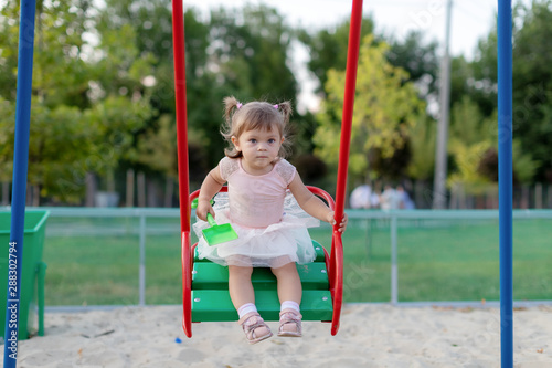 Funny little child, adorable preschooler girl in pretty dress having fun on a swing in the park on summer day