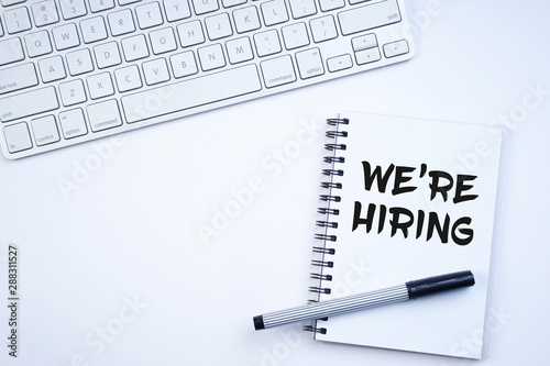 Spiral notebook with "We're hiring" text on keyboard on white background. Recruitment concept. Top view or flat lay.