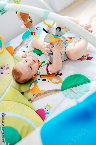 Top view of playful Caucasian chubby six months old baby boy in colorful bodysuit playing with crib toys.