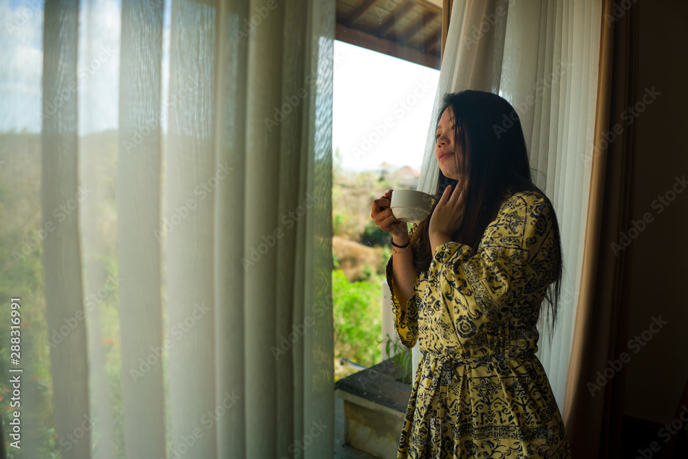 lifestyle portrait of young beautiful happy and relaxed Asian Korean woman in stylish bathrobe by luxury hotel room window having morning coffee enjoying the tropical view