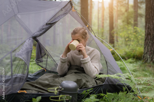 Woman covered with warm sleeping bag meets cold morning in sitting in a touristic camping tent with a cup of hot tea.