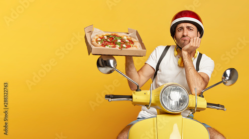 Tablou pe pânză Photo of serious dissatisfied busy pizza man shows tasty junk food on cardboard box, poses on fast transport, wears helmet and white t shirt, being tired of transporting food for customers