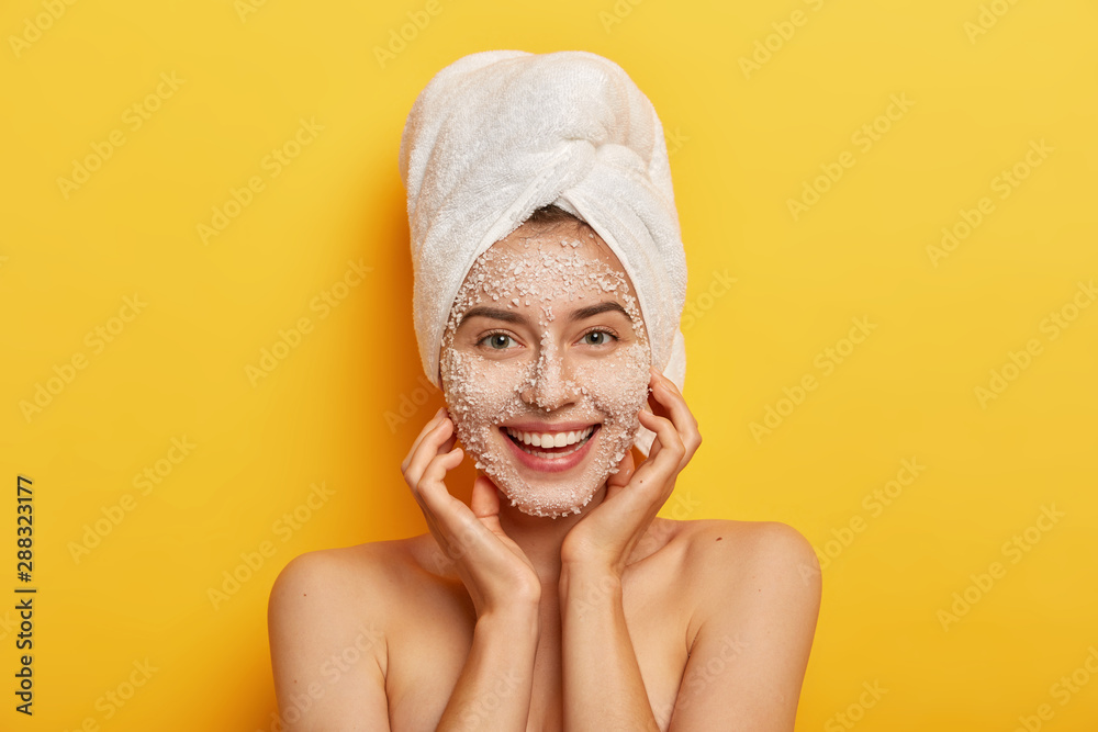 Pleasant looking happy woman unclogges pores, makes beauty step to improve skin, wears nourishing facial scrub, supples complexion, keeps hands on cheeks, stands naked against yellow studio wall Pleasant looking happy woman unclogges pores, makes beauty step to improve skin, wears nourishing facial scrub, supples complexion, keeps hands on cheeks, stands naked against yellow studio wall