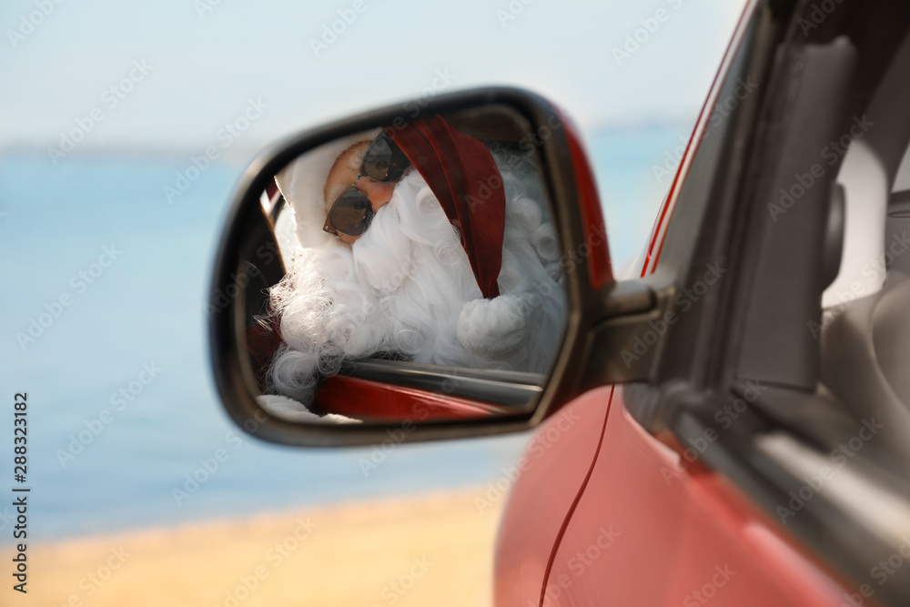Authentic Santa Claus looking into side view mirror of car near sea
