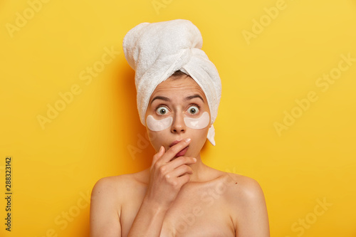 Impressive woman has natural beauty, stares with bugged eyes, applies hydrogel patches which make visible difference to her skin, excess oil production, touches mouth with hands, poses topless