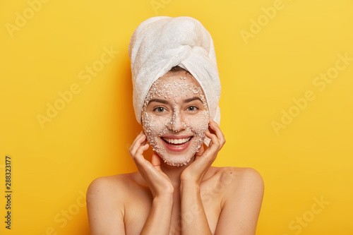 Pleasant looking happy woman unclogges pores, makes beauty step to improve skin, wears nourishing facial scrub, supples complexion, keeps hands on cheeks, stands naked against yellow studio wall
