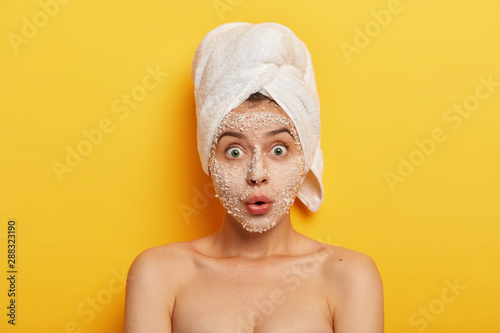 Stupefied young woman applies natural sea salt mask, reduces acnes and pimples, stares with widely opened eyes, healthy skin, cares about complexion, has spa therapy in bathroom. Hygiene and wellness photo