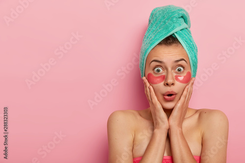 Beautiful shocked lady touches cheeks with both hands  has stupefied look at camera  applies collagen pink patches for reducing fine lines under eyes  has wet hair wrapped in towel  models indoor