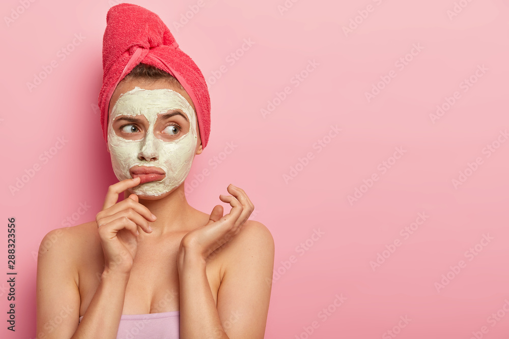Sad upset woman wears white natural mask, has thoughtful expression, wants to have glow perfect smooth skin, applies facial clay mask, towel on head, poses over pink wall with copy space area Sad upset woman wears white natural mask, has thoughtful expression, wants to have glow perfect smooth skin, applies facial clay mask, towel on head, poses over pink wall with copy space area
