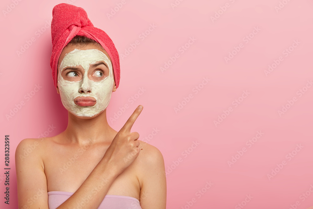 Beauty, spa, treatment concept. Unhappy dissatisfied woman purses lips, applies clay mask for rejuvenation, wrapped in bath towel, points on copy space against pink background, wants fast result