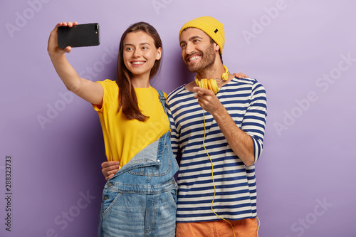 Happy female and male teenagers take selfie on smartphone, smile and embrace, cuddle each other, dressed in fashionable clothes, stand against purple background, point at display, photograph themself photo