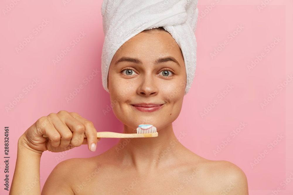 Close up portrait of smiling optimistic woman with fresh skin, holds toothbrush with toothpaste, wears white towel on head, looks directly at camera, has oral hygiene procedure, isolated on pink wall Close up portrait of smiling optimistic woman with fresh skin, holds toothbrush with toothpaste, wears white towel on head, looks directly at camera, has oral hygiene procedure, isolated on pink wall