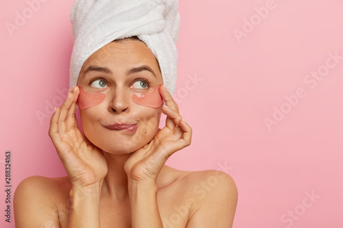 Headshot of attractive young woman applies hydrogel patches under eyes, bites lower lip, removes dark circles, looks aside, stands naked against pink background. Skin care and beauty concept.