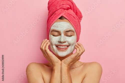 Spa girl applies clay mask on face, keeps eyes closed, touches cheeks, gets pleasure from beauty procedure, refreshes skin, smiles positively isolated on pink background. Relaxation, healthy lifestyle