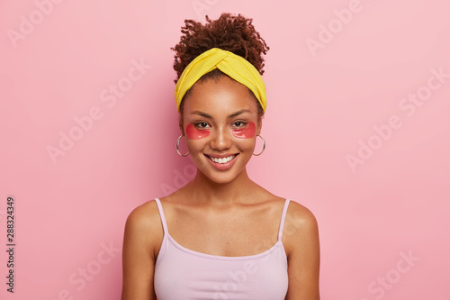 Slika na platnu Portrait of good looking dark skinned young African American woman wears cosmetic collagen patches, wears headband and casual t shirt, isolated over pink background