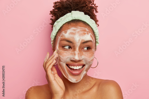 Headshot of pretty dark skinned young model touches cheek with foam, washes face with water and soap, looks gladfully aside, wears round earrings, headband, poses naked against pink background