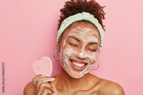 Beautiful optimistic Afro American woman cleanses face with foam, refreshes skin, has well cared complexion, holds heart shaped sponge for beauty procedures, stands bare shoulders with closed eyes photo