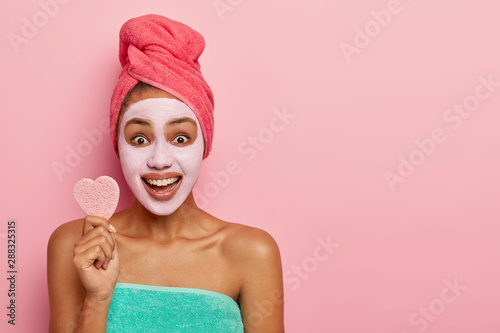 Studio shot of optimistic woman holds soft little sponge for facial treatments, stands wrapped in towel, smiles broadly, applies fresh clay mask for cleaning face, healthy skin. Copy space for text