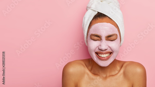 Spa girl with pleased facial expression, applies clay mask on face, gets beauty treatments, wears white soft towel on head, stands with bare shoulders, isolated over pink wall with copy space area