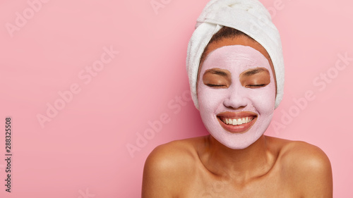 Close up portrait of young female model applies homemade facial clay mask, has white towel wrapped around head, keeps eyes shut, smiles happily, models against pink background. Beauty treatment photo