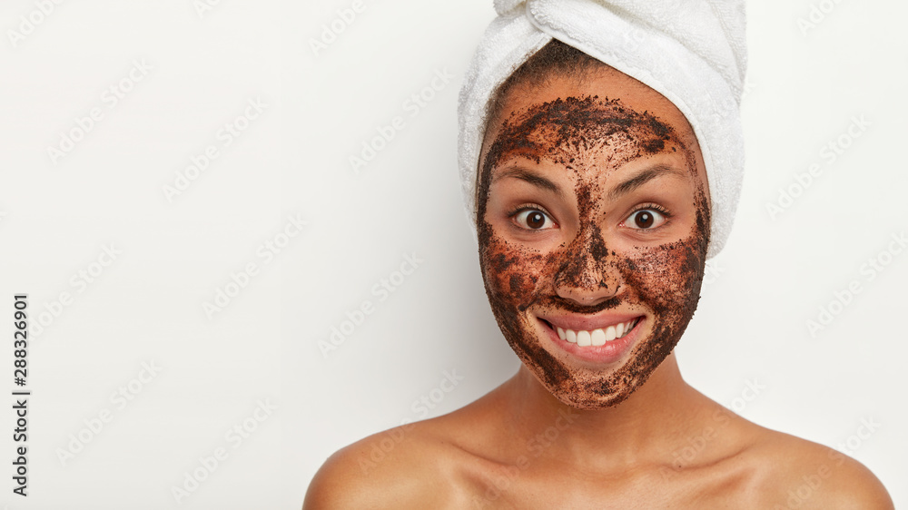 Smiling glad woman with dark skin, refreshes her complexion with coffee scrub mask, looks with widely opened eyes at camera, bites lips and shows white teeth, stands indoor. Body care and beauty Smiling glad woman with dark skin, refreshes her complexion with coffee scrub mask, looks with widely opened eyes at camera, bites lips and shows white teeth, stands indoor. Body care and beauty