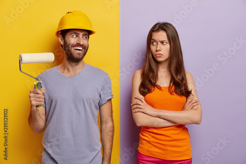 Happy hard working male in hardhat, holds paint roller, displeased wife stands crossed hands and looks at him, do house repairing at home, dressed in casual wear, isolated over two colored background photo