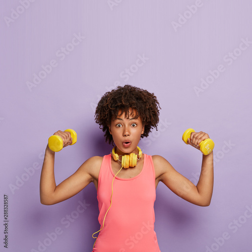 Weight loss and exercising concept. Surprised dark skinned woman with curly hair, raises dumbbells, trains muscles, has easy biceps exercise, wears casual pink top, uses headphones, isolated on purple