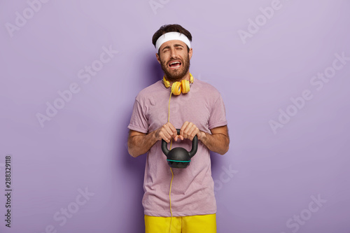 Hard training concept. Sorrowful unhappy young man lifts heavy weight, being demotivated for sport, has no wish to work in gym, wears white headband, casual clothes, listens music during workout photo