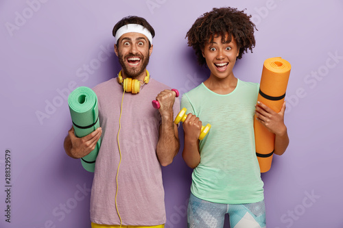 Workout, fitness and sport concept. Cheerful mixed race couple have workout, raise arms with dumbbells, hold karemats, have training in gym. Sporty family go in for sport together. Healthy lifestyle photo