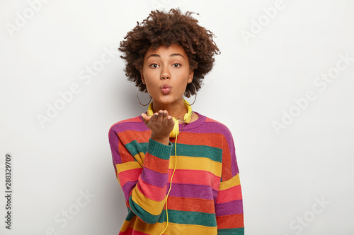 Portrait of beautiful affectionate curly teenage girl keeps palms stretched forward, lips rounded, sends air kiss, wears casual colorful jumper, uses headphones for listening favourite melodies