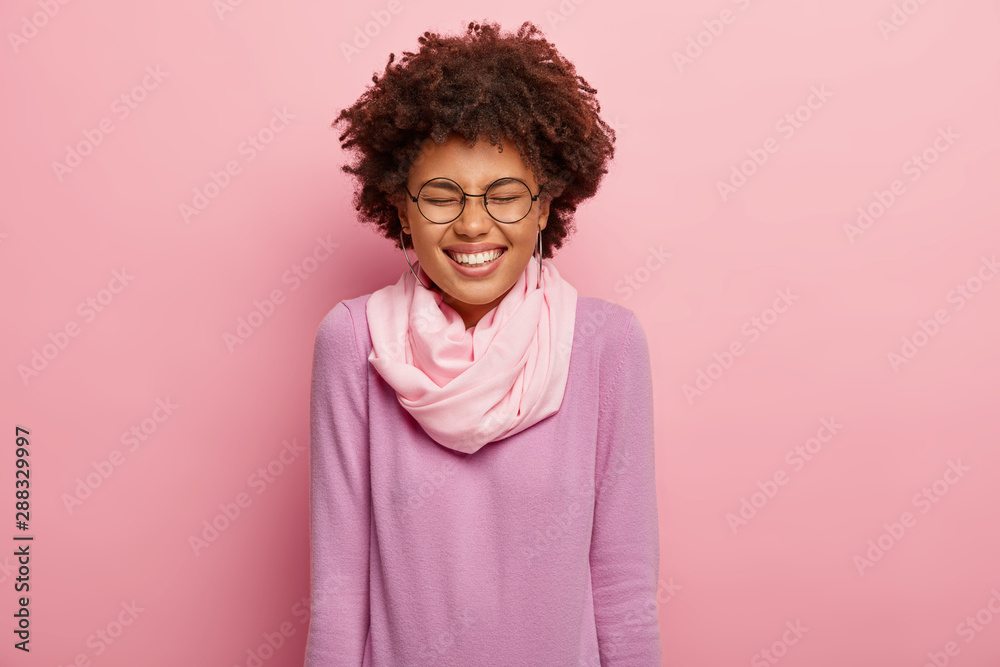 Portrait of cheerful overjoyed young woman with toothy smile, keeps eyes closed, laughs at something funny, has perfect mood, shows white teeth, wears casual jumper and scarf, models indoor. Portrait of cheerful overjoyed young woman with toothy smile, keeps eyes closed, laughs at something funny, has perfect mood, shows white teeth, wears casual jumper and scarf, models indoor.
