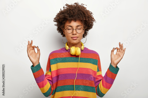 Relaxed beautiful lady with Afro hairstyle meditates indoor, keeps eyes closed, feels satisfied lo listen pleasant music in headphones, wears spectacles, earrings, striped jumper. Body language