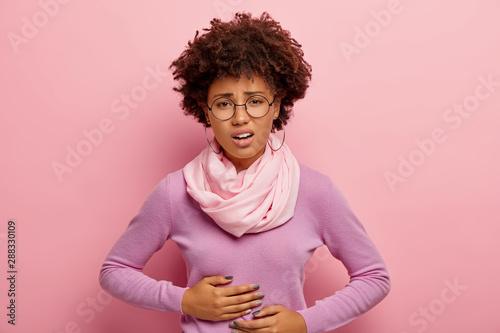 Fototapeta Young Afro American woman suffers from painful cramps in belly, has stomachache,