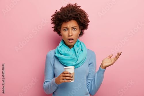 Displeased indignant dark skinned woman with curly hairstyle, raises hand, looks with frustration, dressed in casual blue clothes, holds takeout coffee isolated over pink background. Negative emotions
