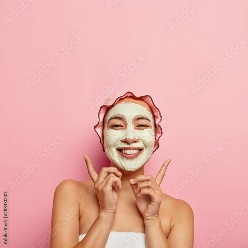 Fototapeta Naklejka Na Ścianę i Meble -  Skin treatment and wellness concept. Cheerful mixed race woman with clay mask of facial cream, points above index fingers, glad to feel fresh, demonstrates copy space for your promotional content.