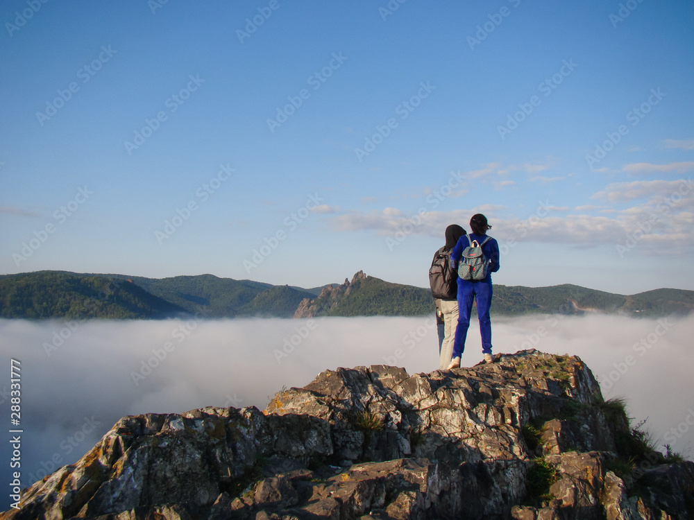 Young people on a rock look into the distance. Around the dense fog. Back view. Concept of mountain tourism, hiking.
