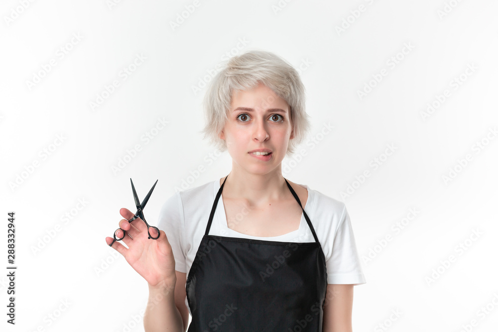 Disturbed young hairdresser in white tee and black apron holding black scissors ready for work standing isolated over white background. Wellness and beauty industry and professional career. Disturbed young hairdresser in white tee and black apron holding black scissors ready for work standing isolated over white background. Wellness and beauty industry and professional career.