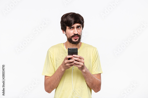 Handsome brunet bearded man with whiskers in a yellow tee holding smartphone expressing emotion of distrust isolated over white background. photo