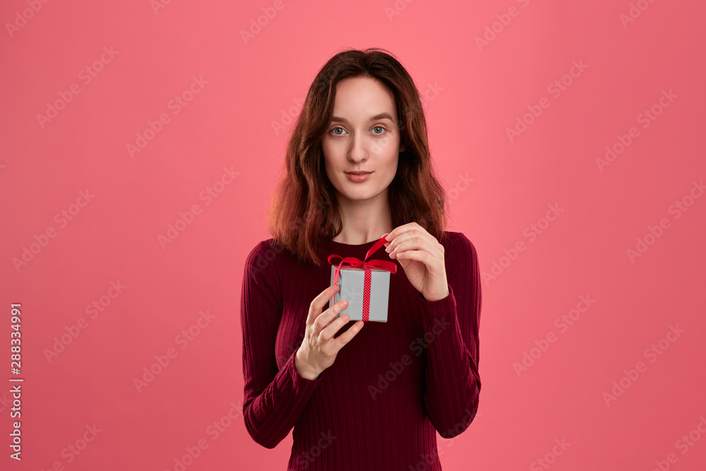 Excited pretty brunette girl opens gift packed in present box with a ribbon standing isolated on a dark pink background and smiling at the camera. Celebrating special event. Excited pretty brunette girl opens gift packed in present box with a ribbon standing isolated on a dark pink background and smiling at the camera. Celebrating special event.