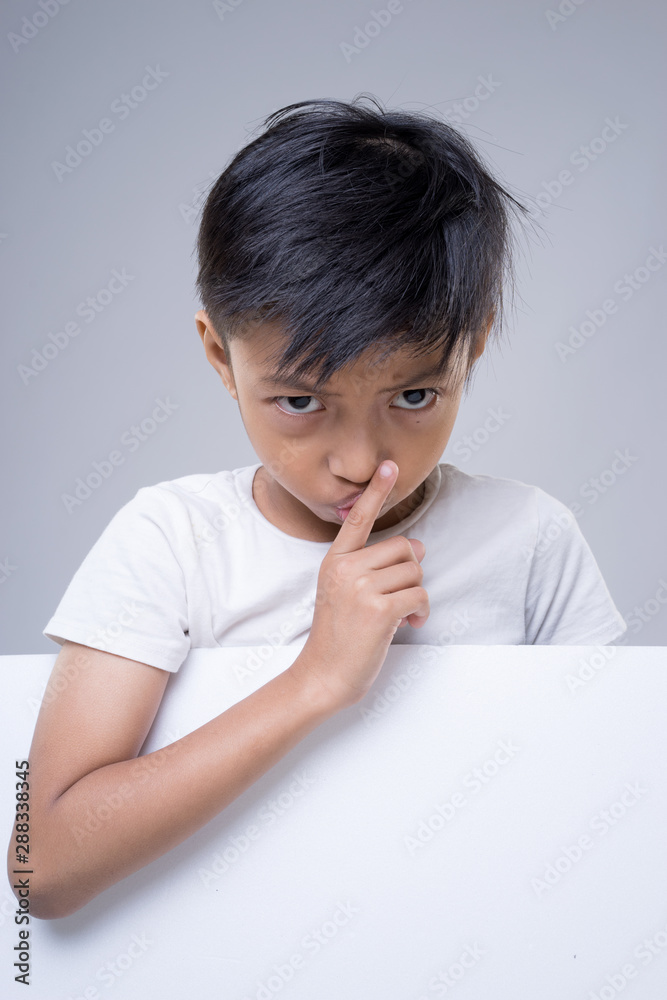 An Asian child wearing white t-shirt putting a finger on his lips a sign of quiet please on grey background. An Asian child wearing white t-shirt putting a finger on his lips a sign of quiet please on grey background.