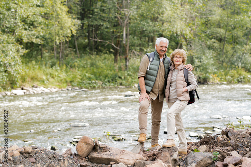 Affectionate mature spouses with backpacks standing on stones on river bank
