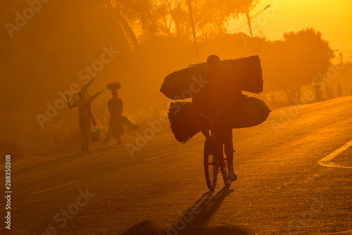 A Mozambican cyclist carrying large bags of coal to market at sunrise in the morning, along a tarmac road. Other commuters carrying items in the background. Nampula Town, Mozambique