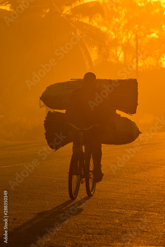 A Mozambican cyclist carrying large bags of coal to market at sunrise in the morning, along a tarmac road. Nampula Town, Mozambique