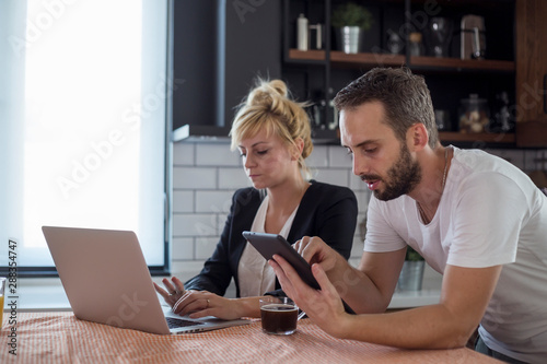 Couple in kitchen in the morning. They are working on tablet and laptop.