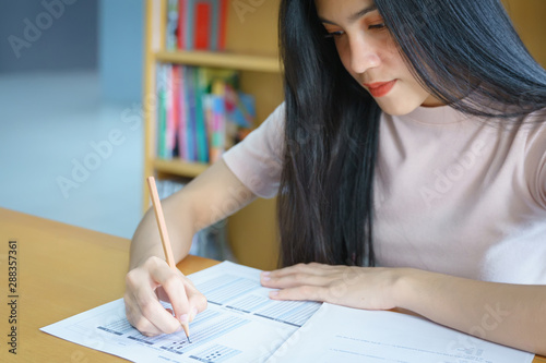 Young female university student concentrate doing language practice examination inside library. Girl student writes the exercise of the examinations. University examination and education concept.