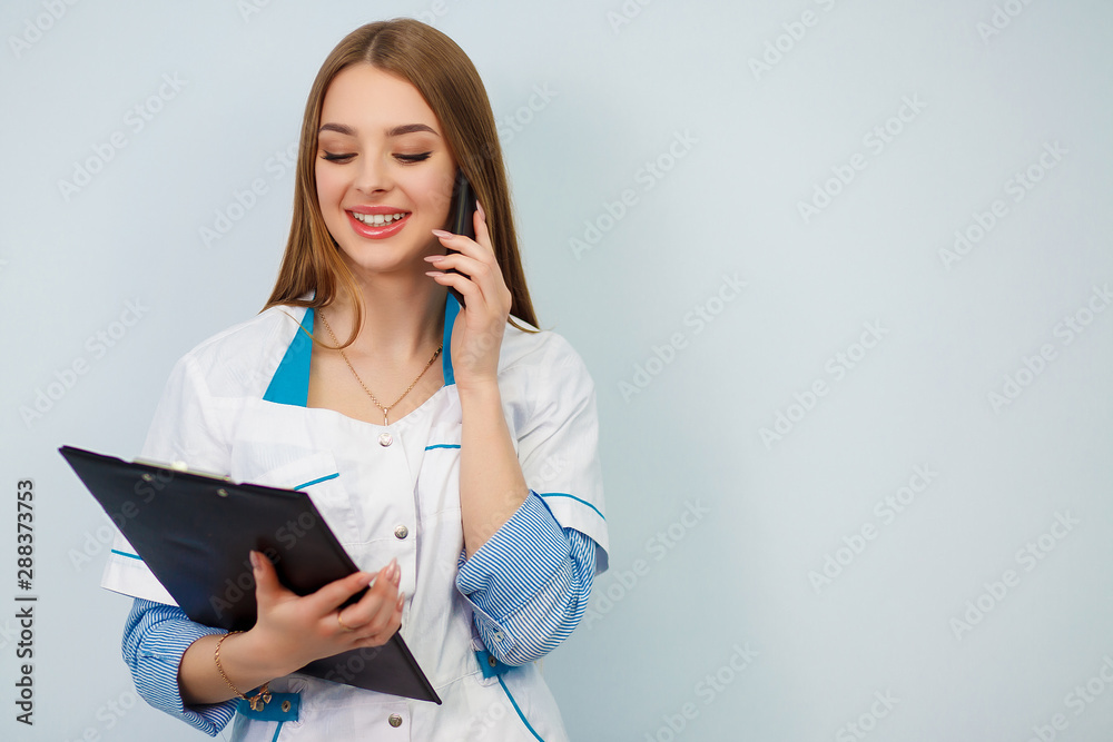 Health care concept. Beautiful girl in a white coat. Copy space. Photo of a young woman doctor posing isolated over white wall background holding clipboard talking by mobile phone Health care concept. Beautiful girl in a white coat. Copy space. Photo of a young woman doctor posing isolated over white wall background holding clipboard talking by mobile phone