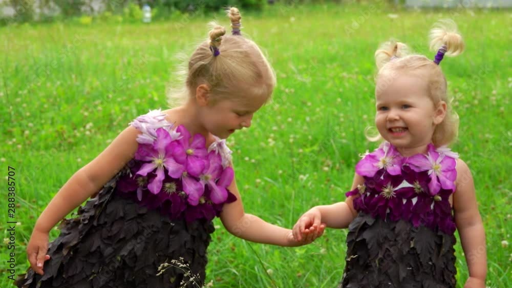 Sisters hold hands, smile and dance. Girls wear unusual dresses made of leaves and flowers Sisters hold hands, smile and dance. Girls wear unusual dresses made of leaves and flowers