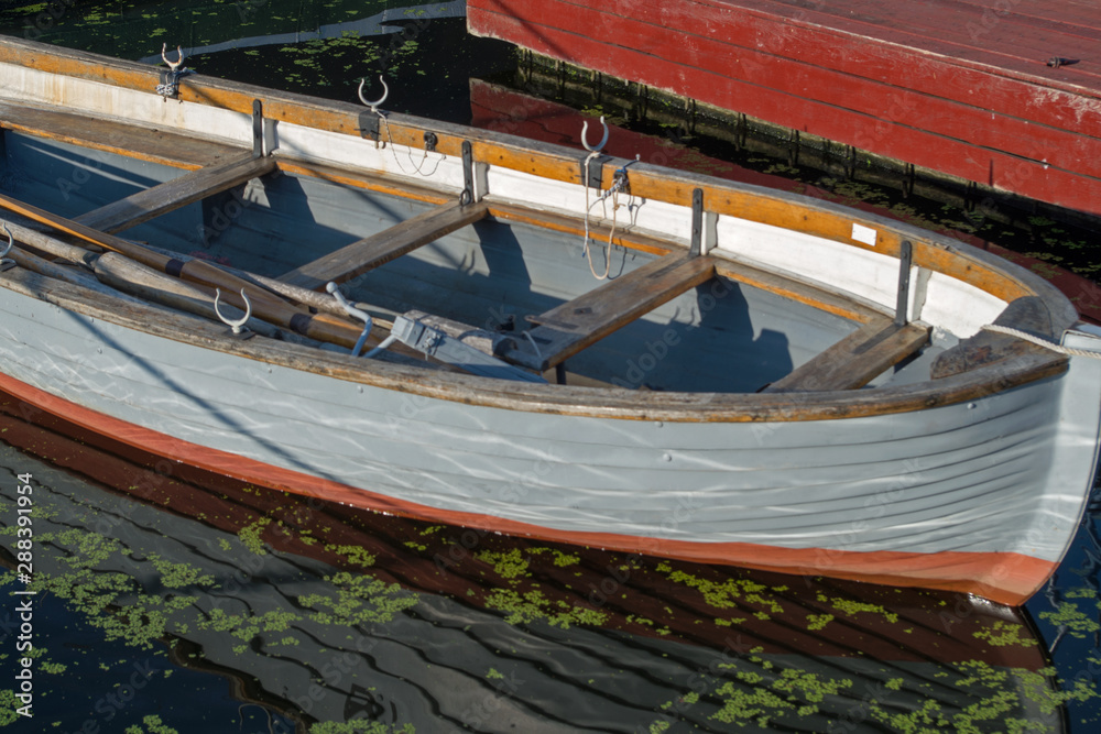 Wooden boat on the pier has a gray side with a number of rowlocks and wooden oars in it, located nose to the viewer at the red pier Wooden boat on the pier has a gray side with a number of rowlocks and wooden oars in it, located nose to the viewer at the red pier