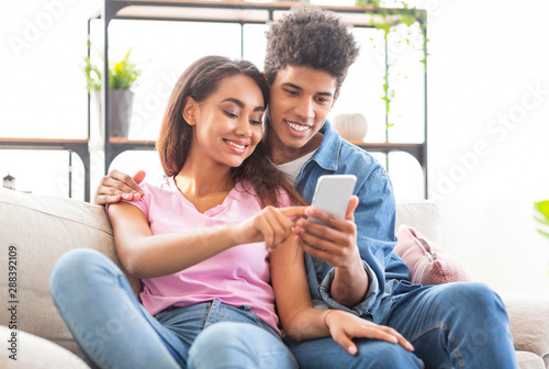 Smiling teenagers scrolling photos in smartphone together sitting at home