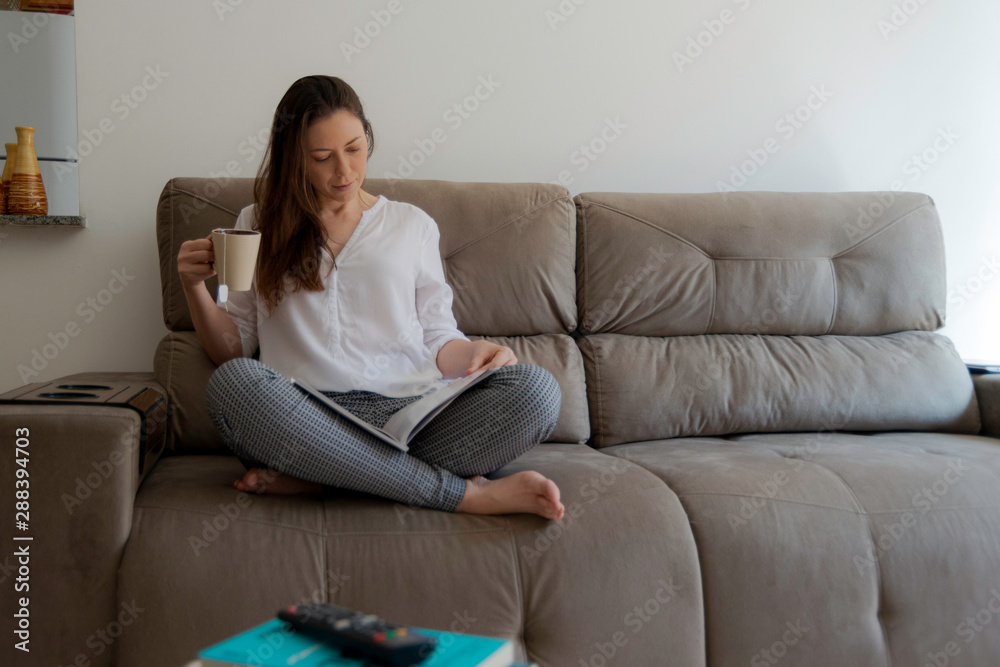 Beautiful woman sitting on the couch reading magazine and having a tea.